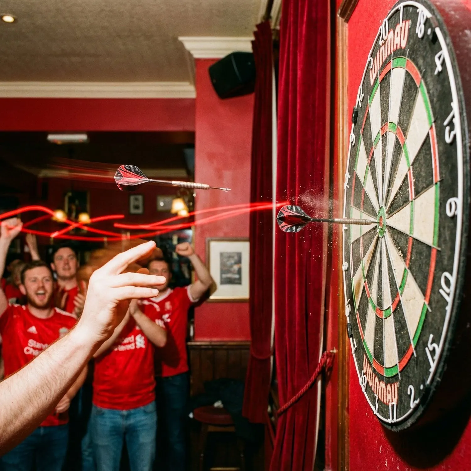 A dart flies toward a board in a red pub while fans cheer behind the thrower.